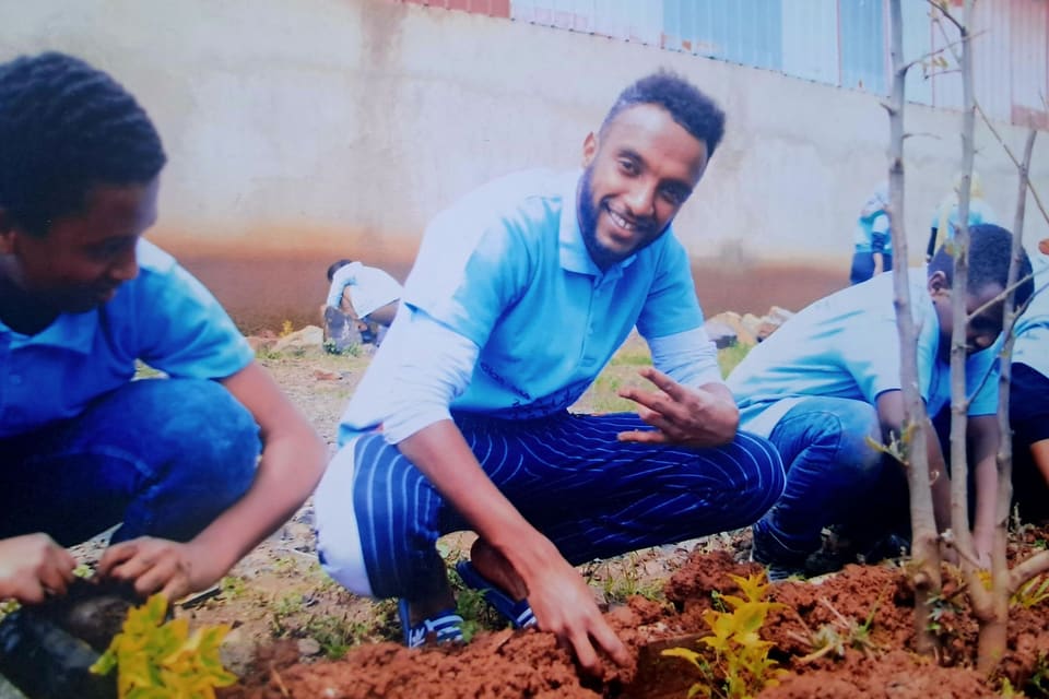 A colleague kneeling in red earth, smiling at the camera while firming soil around a sapling.